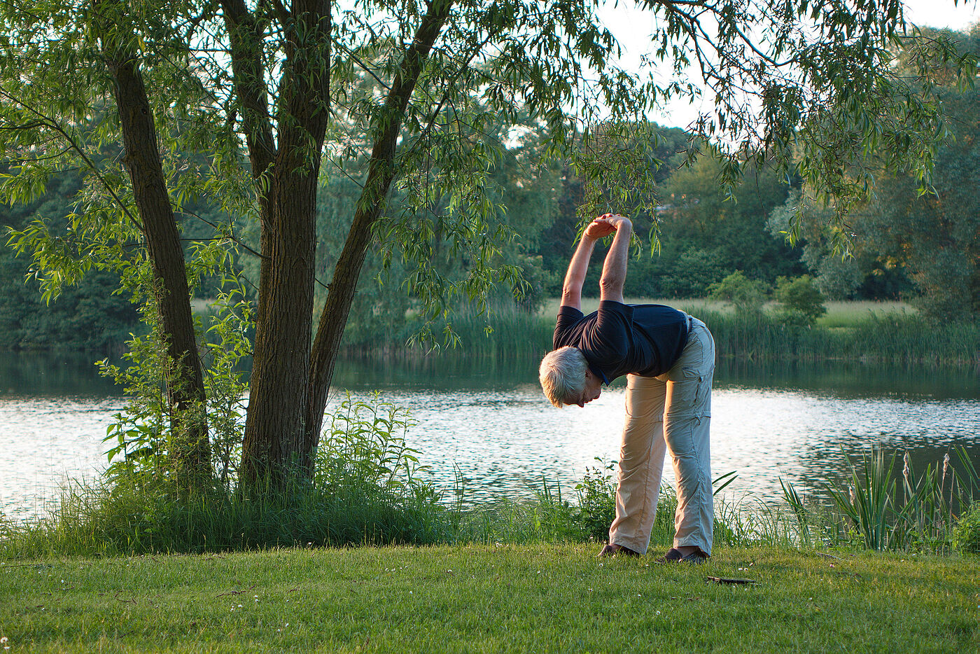 Ein Mann macht Rückenübungen auf einer Grünfläche an einem See.