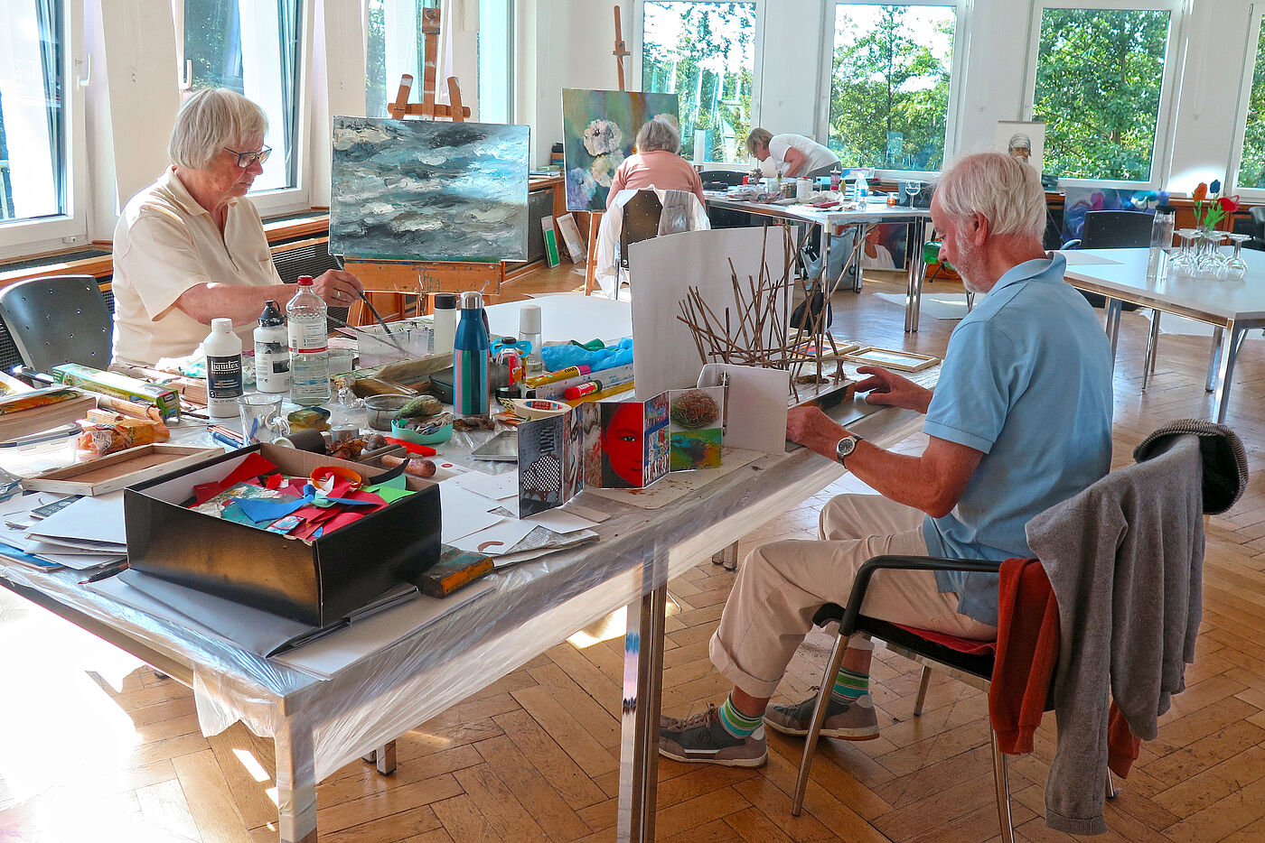 Vier ältere Menschen sitzen in einem Atelier mit Farben, Pinseln und Staffeleien.